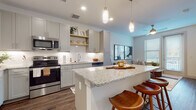 This thoughtfully designed single-floor residence combines modern aesthetics with functional living. The open-concept kitchen is a standout feature, showcasing soft gray shaker-style cabinetry, granite countertops, a full suite of stainless steel appliances, and a generous island with bar seating illuminated by elegant pendant lighting. Warm hardwood-style flooring runs throughout the main living areas, complementing the bright, neutral color palette. Open shelving adds a contemporary touch to the kitchen's design. The kitchen flows seamlessly into the living area, which is bathed in natural light from multiple windows and features a ceiling fan overhead. The bedrooms offer a calm, restful atmosphere with plush carpeting, large windows, and ample natural light. The primary bedroom connects to a well-sized walk-in closet and an en-suite bathroom finished with gray cabinetry matching the kitchen, granite countertops, rich hardwood-style flooring, and large-format marble-style tile in the shower surround. The overall ambiance of the space is clean, contemporary, and cohesive, with consistent finishes and a neutral tone throughout that creates an inviting and polished living environment.