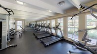 This fitness center is a spacious, single-floor facility designed to accommodate a comprehensive range of workout needs. The interior features a clean, neutral color palette with warm beige walls, white beam-detailed ceilings, and dark rubber flooring that runs throughout the entire space, providing both durability and a professional aesthetic.

The layout is elongated, offering generous floor space organized into distinct functional zones. Along one side, a row of treadmills and elliptical machines lines the wall of large windows, which flood the space with natural light and offer views of an outdoor pool area, creating an engaging environment for cardio workouts. A cable machine and multi-function strength equipment are positioned centrally, complemented by a dedicated free weight area featuring a tiered dumbbell rack and an adjustable weight bench.

Exposed beam detailing on the ceiling adds architectural character, while flush-mounted light fixtures provide consistent overhead illumination throughout. Multiple access doors, including an emergency exit, contribute to practical circulation within the space. A water cooler station is conveniently positioned near the equipment. Mirrors along portions of the walls further enhance the sense of openness and functionality throughout this well-appointed fitness facility.