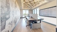 Located at 360 Acoma Street in Denver, Colorado, this compact yet thoughtfully appointed study offers a distinctive and inspiring workspace environment. The room is anchored by a set of long, shared workstations fitted with acoustic privacy panels and ergonomic rolling chairs, providing a focused and functional co-working setup.

One of the most striking design elements is the large-scale feature wall adorned with an intricate mural composed of vintage scientific and exploratory illustrations — including anatomical skeletons, celestial instruments, telescopes, hot air balloons, and botanical imagery — all set against a typographic background. The artwork lends the room a deeply intellectual and creative character.

Overhead, a bold geometric ceiling installation features a lattice of brass-toned rods fitted with exposed Edison-style bulbs, creating warm, ambient lighting with an industrial-meets-artisan aesthetic. The light hardwood flooring complements the neutral wall tones and adds warmth to the space.

Floor-to-ceiling windows along one side of the room flood the interior with natural light and frame open outdoor views, enhancing the sense of space and connection to the surrounding environment. A wall-mounted whiteboard provides a collaborative surface for notes and planning. Together, these elements make this study a functional, visually rich, and productive workspace.