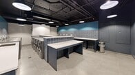 This thoughtfully designed laundry facility offers a comfortable and functional experience across a single, open floor plan. The space features an industrial-chic aesthetic, with exposed black ductwork and piping running along a dark ceiling, complemented by sleek round pendant lights that provide ample illumination throughout.

The main laundry area is equipped with a row of stainless steel washers and dryers, accompanied by generously sized folding counters finished in white with navy blue tile bases. A teal accent wall with subway tile detailing adds a vibrant pop of color, while a utility sink and informational signage enhance the room's practicality. Large-format light grey floor tiles run throughout, creating a clean, cohesive look.

A distinctive "Lost Socks" wall with individual hooks offers a clever and functional touch for misplaced items. Unique newspaper-print wallcovering lines portions of the walls, adding a creative, editorial character to the space.

A separate lounge area provides residents with a comfortable place to wait, furnished with a blue sofa, a round dark table, and curved upholstered chairs. Framed artwork adorns the textured walls, elevating the overall ambiance. The combination of functional amenities and thoughtful design details makes this a standout communal facility.
