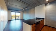 This one-bedroom, one-bathroom unit showcases a compelling industrial-modern design that blends raw architectural elements with contemporary finishes. Throughout the living areas, rich dark hardwood flooring contrasts with exposed concrete ceilings, which are fitted with track lighting that provides both functional illumination and an urban aesthetic. Conduit runs visibly along the walls, reinforcing the loft-inspired character of the space.

The open-concept kitchen features a central island with a dark granite countertop, an undermount sink, and warm-toned wood cabinetry—including a distinctive wine rack detail above. Stainless steel appliances, including a refrigerator, range, and microwave, complete the kitchen. The island flows naturally into the living area, which benefits from a sliding glass door that opens to a private balcony overlooking a tree-lined outdoor view, filling the space with natural light.

The bedroom offers carpeted flooring for comfort, an exposed concrete ceiling, and a reach-in closet providing ample storage. The bathroom is finished with matching dark granite countertops, warm wood vanity cabinetry, a large mirror, and a tub-shower combination tiled in classic grey subway tile, all set atop hardwood-style flooring.