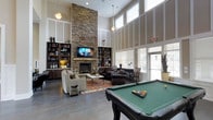This impressive single-floor lounge space commands attention with its soaring double-height ceilings and an abundance of natural light streaming through large, floor-to-ceiling windows. The centerpiece of the room is a dramatic stacked-stone fireplace that rises to full ceiling height, flanked by built-in bookshelves that provide both storage and visual symmetry. A flat-screen television is mounted above the fireplace mantle, making it a natural focal point for the comfortable seating arrangement below, which includes plush armchairs and dark leather sofas arranged around a coffee table atop a warm area rug. Pendant lighting and suspended fixtures complement the room's vertical scale, adding warmth to the open layout. In the foreground, a full-size pool table with a green felt surface and racked balls invites recreation and social interaction. The polished concrete flooring ties the space together, offering a sleek contrast to the rustic stone feature wall. Large windows with blinds line the perimeter, offering views to the exterior while maintaining privacy. The overall ambiance blends comfortable, residential warmth with an amenity-rich, communal character — ideal for gathering, relaxing, or recreation.