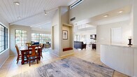 Located at 8115 N 18th St in Phoenix, Arizona, this single-floor space offers a well-designed, open layout that maximizes comfort and functionality. The interior is immediately distinguished by its soaring vaulted ceilings with exposed white-painted beams, which lend an airy, open feel throughout. Light-toned ceramic tile flooring runs consistently across all areas, contributing to a cohesive and clean aesthetic.

The living room is a spacious and inviting area, featuring large floor-to-ceiling grid-style windows that flood the room with natural light and provide views of the lush outdoor surroundings, including palm trees and a pool area. A ceiling fan adds to the comfort of the space. A dedicated sitting nook with built-in wall shelving serves as a charming reading corner, bathed in natural light from an adjacent window.

The dining area is anchored by a substantial wooden dining table and is framed by the same expansive grid windows, creating a bright and welcoming atmosphere for gatherings. A central dividing column with a built-in display niche adds architectural character and thoughtfully delineates the open living and dining zones. The overall ambiance is warm, light-filled, and thoughtfully laid out, making this Phoenix space both functional and visually appealing.