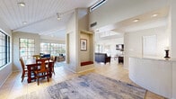 Located at 8115 N 18th St in Phoenix, Arizona, this single-floor space offers a well-designed, open layout that maximizes comfort and functionality. The interior is immediately distinguished by its soaring vaulted ceilings with exposed white-painted beams, which lend an airy, open feel throughout. Light-toned ceramic tile flooring runs consistently across all areas, contributing to a cohesive and clean aesthetic.

The living room is a spacious and inviting area, featuring large floor-to-ceiling grid-style windows that flood the room with natural light and provide views of the lush outdoor surroundings, including palm trees and a pool area. A ceiling fan adds to the comfort of the space. A dedicated sitting nook with built-in wall shelving serves as a charming reading corner, bathed in natural light from an adjacent window.

The dining area is anchored by a substantial wooden dining table and is framed by the same expansive grid windows, creating a bright and welcoming atmosphere for gatherings. A central dividing column with a built-in display niche adds architectural character and thoughtfully delineates the open living and dining zones. The overall ambiance is warm, light-filled, and thoughtfully laid out, making this Phoenix space both functional and visually appealing.