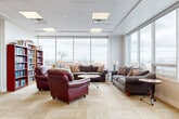 Located at 725 Highway 15 in Kingston, Ontario, this generously sized social room is a thoughtfully designed communal space that serves as a hub for gathering and relaxation. The room is spread across a single floor and is divided into several distinct functional zones that flow seamlessly into one another.

The lounge area is furnished with rich burgundy leather armchairs and plush grey sofas arranged around a central coffee table, creating a welcoming and comfortable setting for conversation. Floor-to-ceiling windows line the exterior walls, flooding the space with abundant natural light and offering sweeping panoramic views of the surrounding Kingston skyline and landscape.

Adjacent to the lounge is a formal dining area featuring a solid wood table with matching cushioned chairs, ideal for group meals or meetings. A well-stocked library corner with multiple tall bookshelves adds an intellectual and cozy character to the room, complemented by a large decorative plant and wall art nearby.

The kitchenette is equipped with warm wood cabinetry, a stainless steel refrigerator, microwave, and a breakfast bar with bar stools — providing full functionality for catered events or casual use. Throughout the space, patterned carpet tiles in neutral tones and a drop-panel ceiling with recessed lighting contribute to a clean, modern, and inviting ambiance.