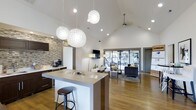 Located at 2223 East 151st Street in Carmel, Indiana, the Carmel Landing clubhouse is a single-floor community space thoughtfully designed to serve residents with both style and functionality. The open-concept interior is anchored by soaring vaulted ceilings that create an expansive, airy atmosphere throughout. Warm hardwood flooring runs continuously across the space, complementing the modern aesthetic.

The kitchenette area features a large central island with bar seating, pendant globe lighting overhead, and dark cabinetry accented by a striking stacked stone accent wall. A coffee station adds a welcoming, residential touch. Recessed lighting and decorative pendants provide warm, layered illumination throughout.

The adjacent lounge area is furnished with comfortable seating, a wall-mounted flat-screen television, and a ceiling fan, making it ideal for casual gatherings and community events. Floor-to-ceiling glass doors and windows flood the interior with natural light while offering a direct view into the fully equipped fitness center, which is visible from the main common area and features cardio and exercise equipment. The overall ambiance is contemporary and inviting, blending clean lines, natural textures, and modern finishes into a cohesive and well-appointed community hub.