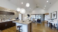 Located at 2223 East 151st Street in Carmel, Indiana, the Carmel Landing clubhouse is a single-floor community space thoughtfully designed to serve residents with both style and functionality. The open-concept interior is anchored by soaring vaulted ceilings that create an expansive, airy atmosphere throughout. Warm hardwood flooring runs continuously across the space, complementing the modern aesthetic.

The kitchenette area features a large central island with bar seating, pendant globe lighting overhead, and dark cabinetry accented by a striking stacked stone accent wall. A coffee station adds a welcoming, residential touch. Recessed lighting and decorative pendants provide warm, layered illumination throughout.

The adjacent lounge area is furnished with comfortable seating, a wall-mounted flat-screen television, and a ceiling fan, making it ideal for casual gatherings and community events. Floor-to-ceiling glass doors and windows flood the interior with natural light while offering a direct view into the fully equipped fitness center, which is visible from the main common area and features cardio and exercise equipment. The overall ambiance is contemporary and inviting, blending clean lines, natural textures, and modern finishes into a cohesive and well-appointed community hub.