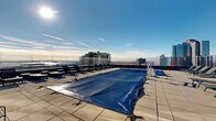 Located atop a modern high-rise at 10-14 LeCount Place in New Rochelle, NY, this generously sized rooftop terrace offers an exceptional outdoor amenity experience. The space is anchored by a large swimming pool, currently covered, which serves as the central focal point of the deck. Surrounding the pool is a spacious tiled terrace outfitted with numerous chaise lounges, providing ample room for sunbathing and relaxation.

The rooftop is enclosed by sleek glass panel railings that preserve unobstructed, panoramic views in every direction — from the New Rochelle and greater New York City skyline to the shimmering expanse of Long Island Sound and its islands beyond. String lights are strung across portions of the deck, adding a warm, ambient atmosphere for evening use.

The adjacent building facade is clad in a contemporary curtain wall of glass and steel, with direct access points leading into the interior. Planters with greenery are positioned throughout the terrace, softening the urban aesthetic. The open-sky exposure ensures abundant natural light throughout the day, making this rooftop an elevated retreat that combines modern design with spectacular outdoor living above the heart of New Rochelle.
