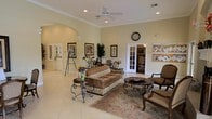 This single-story space serves as a well-appointed lobby or common area, designed to create a comfortable and welcoming environment. The room features smooth, light-colored walls accented by classic crown molding and arched architectural details that lend an elegant touch. Large French doors with glass panels allow natural light to filter through while providing a sense of openness and connectivity to adjacent spaces.

The flooring consists of light-toned tile throughout, offering a clean and polished look. A ceiling fan with recessed lighting overhead ensures comfort and ample illumination across the space. The furnishings are traditionally styled, including a patterned sofa, ornate wrought-iron accent tables, and upholstered armchairs, all arranged atop a decorative area rug to define the seating area.

Decorative elements include framed artwork, potted plants, and fresh floral arrangements that add warmth and color. A wall clock and a community bulletin board are visible, suggesting a shared or communal setting. The overall ambiance is refined yet approachable, blending classic design details with practical, functional elements suited for a gathering or reception space.