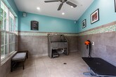 Located in Alpharetta, Georgia, this single-story pet grooming facility is designed with both functionality and comfort in mind. The space features a consistent and inviting aesthetic throughout, characterized by soft aqua-painted upper walls and large-format neutral tile cladding the lower walls, separated by a decorative mosaic tile border that adds a refined visual detail. Light-colored tile flooring runs throughout, making the space easy to clean and maintain.

The grooming area is equipped with a professional-grade stainless steel pet bathing tub, complete with built-in steps for easy pet access and a floor drain for efficient water management. A wall-mounted high-velocity pet dryer is conveniently positioned nearby, and an adjustable grooming table with paw-print detailing provides a dedicated space for finishing work. Framed photos of pets and their owners decorate the walls, contributing to a warm and welcoming atmosphere.

Natural light enters through multiple large windows, offering views of surrounding greenery and keeping the space bright. A glass-paned exterior door provides additional light and direct outdoor access. A padded bench along one window wall offers a comfortable waiting area. The space is also equipped with practical amenities including paper towel dispensers and a stainless steel waste bin.