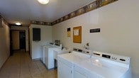 This shared laundry facility offers a practical and functional space designed to serve multiple users efficiently. The room is outfitted with several top-load washing machines and front-facing dryers arranged along the walls, providing ample capacity for regular use. Light-colored walls and tile flooring contribute to a clean, utilitarian aesthetic, while a decorative border running along the upper perimeter of the room adds a modest touch of character. Overhead lighting ensures the space is well-lit throughout. A cork bulletin board is mounted on the wall, displaying notices and informational postings for users, alongside posted reminders such as lint removal instructions — a detail that speaks to the room's emphasis on shared responsibility and upkeep. A coin-operated or card-based payment mechanism is visible on the machines, indicating a self-service setup. The layout is straightforward and accessible, with enough open floor space to move comfortably while loading and unloading laundry. Overall, this laundry room is a well-organized, no-frills communal amenity built for reliability and ease of use.