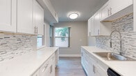 This single-floor, two-bedroom, one-bathroom residence offers a thoughtfully updated interior with modern finishes across a comfortable footprint. The galley-style kitchen is a standout feature, showcasing white shaker cabinetry with brushed nickel hardware, quartz countertops, a mosaic tile backsplash in neutral tones, and stainless steel appliances. A window at the end of the kitchen brings in natural light and views of surrounding greenery, enhancing the bright, clean aesthetic.

Wood-look plank flooring runs throughout the unit, providing a cohesive and durable surface in every room. The living area is generously sized, featuring a ceiling fan with light fixture and a bold accent wall, adding depth to the otherwise neutral palette. Both bedrooms are well-proportioned, each with windows that frame outdoor views and allow ample natural light.

The bathroom includes a white shaker vanity with a dark countertop, undermount sink, brushed nickel fixtures, and a full tub-shower combination with white tile surround. An adjacent linen closet provides convenient built-in storage. A separate vanity area with additional cabinetry and a mirrored fixture offers extra functionality. The overall design is clean, modern, and move-in ready.