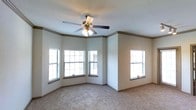 This single-story space showcases a well-appointed interior highlighted by a distinctive angled bay window configuration, allowing natural light to filter in from multiple directions. The main room features neutral-toned carpet flooring that complements the soft, light-colored walls. Crown molding runs along the upper perimeter of the room, adding a refined architectural detail. A ceiling fan with an integrated light fixture is centrally mounted, providing both ventilation and ambient lighting. Multiple windows fitted with horizontal blinds offer privacy while still inviting in daylight. The overall ambiance is clean, open, and move-in ready, with a calm, neutral palette that lends itself well to a variety of interior design styles. The combination of classic trim work and practical features creates a comfortable and functional living environment.