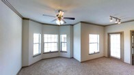 This single-story space showcases a well-appointed interior highlighted by a distinctive angled bay window configuration, allowing natural light to filter in from multiple directions. The main room features neutral-toned carpet flooring that complements the soft, light-colored walls. Crown molding runs along the upper perimeter of the room, adding a refined architectural detail. A ceiling fan with an integrated light fixture is centrally mounted, providing both ventilation and ambient lighting. Multiple windows fitted with horizontal blinds offer privacy while still inviting in daylight. The overall ambiance is clean, open, and move-in ready, with a calm, neutral palette that lends itself well to a variety of interior design styles. The combination of classic trim work and practical features creates a comfortable and functional living environment.