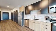 This single-floor unit showcases a clean, contemporary design highlighted by light wood-tone flooring, crisp white walls, and dark trim and door frames that create a striking contrast throughout. The open layout connects the main living area to the kitchen, fostering a spacious and cohesive feel. Recessed lighting illuminates the interiors, complementing the natural light that pours in through large sliding glass doors leading to a private balcony with views of the surrounding neighborhood.

The kitchen is a standout feature, outfitted with a two-tone cabinet system pairing light wood lower cabinets with matte black upper cabinets. A white subway tile backsplash adds texture and visual interest, while light-toned countertops provide ample workspace. Stainless steel appliances — including a refrigerator, electric range, over-the-range microwave, and dishwasher — complete the functional and stylish setup. A matte black faucet and undermount sink add refined detail.

The living areas are generously proportioned, offering flexible room arrangements. Dark-framed interior doors and frosted glass closet panels contribute to the unit's modern aesthetic, while the overall neutral palette creates a versatile, move-in-ready environment.