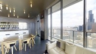 This single-floor space makes a striking first impression with its expansive floor-to-ceiling glass walls that frame panoramic city skyline views, flooding the interior with natural light throughout the day. The open-plan layout seamlessly connects the living and dining areas, creating a cohesive and functional environment. The living area features a plush cream sofa positioned to take full advantage of the dramatic urban vistas beyond the glass, while the dining area is furnished with a light wood table and matching chairs, offering a casual yet refined setting. Dark slate-toned floor tiles run throughout, grounding the space with a sleek, contemporary feel that contrasts beautifully with the lighter furnishings. The design aesthetic blends modern and industrial influences, highlighted by textured stone-effect wall cladding, dark framed glass partitions, and recessed ceiling lighting. A compact kitchen area is visible in the background, neatly integrated into the overall layout. A colorful piece of wall art adds a vibrant focal point, injecting personality into the otherwise neutral palette. The combination of elevated city views, thoughtful design details, and an open, airy atmosphere makes this a distinctive and memorable space.