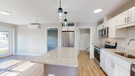 Located at 231 Scarborough Downs Rd in Scarborough, Maine, this thoughtfully designed one-bedroom, one-bathroom residence offers a practical and modern single-floor layout. The kitchen serves as the centerpiece of the home, showcasing white shaker-style cabinetry, light granite countertops, and a contrasting gray central island illuminated by dual pendant lights. Stainless steel appliances — including a refrigerator, range, microwave, and dishwasher — complete this functional and attractive space. Light hardwood-style flooring runs throughout the main living areas, contributing to a bright, cohesive aesthetic. Natural light pours in through windows near the entry door, which features a sidelight panel. A wall-mounted mini-split unit provides climate control. The bedroom offers comfortable carpeted flooring, neutral wall tones, a walk-in closet, and a window with outdoor views, along with a baseboard heating unit. The bathroom is clean and efficiently arranged, featuring a white vanity with brushed nickel hardware, a walk-in shower with white surround, and matching hardwood-style flooring. Overall, the space presents a fresh, modern feel with quality finishes throughout.