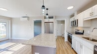 Located at 231 Scarborough Downs Rd in Scarborough, Maine, this thoughtfully designed one-bedroom, one-bathroom residence offers a practical and modern single-floor layout. The kitchen serves as the centerpiece of the home, showcasing white shaker-style cabinetry, light granite countertops, and a contrasting gray central island illuminated by dual pendant lights. Stainless steel appliances — including a refrigerator, range, microwave, and dishwasher — complete this functional and attractive space. Light hardwood-style flooring runs throughout the main living areas, contributing to a bright, cohesive aesthetic. Natural light pours in through windows near the entry door, which features a sidelight panel. A wall-mounted mini-split unit provides climate control. The bedroom offers comfortable carpeted flooring, neutral wall tones, a walk-in closet, and a window with outdoor views, along with a baseboard heating unit. The bathroom is clean and efficiently arranged, featuring a white vanity with brushed nickel hardware, a walk-in shower with white surround, and matching hardwood-style flooring. Overall, the space presents a fresh, modern feel with quality finishes throughout.