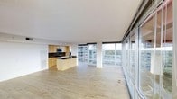 This single-floor unit showcases a seamless open-concept layout that integrates the kitchen, dining, and living areas into one expansive, light-filled space. Light hardwood flooring runs throughout, contributing to a warm yet contemporary aesthetic.

The kitchen is outfitted with light wood cabinetry, dark stone countertops, a dark tile backsplash, and stainless steel appliances including a built-in microwave. A central island provides additional prep and seating space, anchoring the kitchen within the open floor plan.

The most striking feature of the space is the wall of floor-to-ceiling windows along one side, flooding the interior with abundant natural light and offering unobstructed views of the surrounding urban skyline and tree canopy below. A sliding glass door system provides access to the exterior, further enhancing the connection between indoors and outdoors.

The open layout, neutral palette, and expansive glazing combine to create an airy, modern ambiance with a strong sense of elevation and spaciousness throughout.