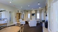 This single-floor space presents a well-appointed communal lounge area designed with comfort and elegance in mind. The room features neutral-toned walls accented by classic crown molding, creating a refined architectural backdrop. Recessed ceiling lights provide consistent, warm illumination throughout the space, complementing the natural light that filters in through multiple large windows dressed with floor-length, neutral-toned draping curtains with coordinating blinds.

The seating area is thoughtfully arranged with plush white upholstered armchairs and sofas positioned around a central coffee table, encouraging conversation and relaxation. A flat-screen television is wall-mounted between the windows, adding a functional entertainment element to the lounge. A substantial wood display cabinet with open shelving stands along one wall, offering both storage and decorative display space.

The flooring transitions between light tile and a rich, dark area rug that anchors the main seating arrangement, adding warmth and definition to the space. Decorative plants and tasteful accessories are placed throughout, enhancing the room's inviting ambiance. A dining area with a round table and chairs is partially visible, suggesting the space serves multiple social functions. Overall, the lounge conveys a clean, classic aesthetic suited for communal use.