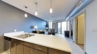 This single-floor space showcases an open-concept layout that seamlessly connects the kitchen and living areas. The kitchen features a generous quartz countertop island with a stainless steel sink, complemented by dark espresso cabinetry with brushed metal hardware. Three cylindrical pendant lights hang above the island, providing focused task lighting with a contemporary aesthetic. An exposed ductwork detail on the ceiling adds an industrial accent to the space.

The living area is defined by a bold slate-blue accent wall, creating a distinct yet cohesive separation from the lighter kitchen zone. Large floor-to-ceiling sliding glass doors flood the space with natural light and provide direct access to an outdoor balcony, offering views of the surrounding landscape. Light wood-toned flooring extends throughout, enhancing the sense of openness.

A hallway visible to the right suggests additional rooms beyond the main living space. The overall ambiance balances modern industrial elements with warm, residential touches, resulting in a functional and stylish environment suited for everyday living and entertaining.