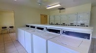 This single-story laundry facility is designed with practicality and efficiency in mind. The room is outfitted with a row of commercial-grade top-loading washing machines — branded Speed Queen — aligned along the front, paired with a corresponding bank of stacked dryers positioned along the back wall. The machines are white and well-maintained, conveying a clean and orderly environment suitable for shared use.

The space is finished with light-toned walls and tile flooring, contributing to a bright and hygienic atmosphere. Overhead, a fluorescent light fixture provides ample illumination throughout the room, while a ceiling fan aids in air circulation, keeping the environment comfortable during use. A ventilation duct is also visible in the ceiling, supporting proper airflow.

The layout is straightforward and functional, with clear access to all machines and enough open floor space to move comfortably between them. A doorway visible at the rear of the room suggests additional access or utility space beyond. Overall, this laundry facility prioritizes convenience and capacity, making it a practical shared amenity.