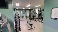 This single-floor fitness room is designed to support a comprehensive workout experience. The space is outfitted with a full dumbbell rack holding a wide range of free weights, providing options for various strength training exercises. A multi-function cable and pulley machine serves as a centerpiece for resistance training, complemented by an adjustable weight bench suitable for a variety of exercises. Floor-to-ceiling mirrors line the walls, visually expanding the room while allowing users to monitor their form during workouts. The flooring features a durable, cushioned surface well-suited for gym use. Overhead fluorescent lighting ensures the space is brightly and evenly illuminated throughout. The walls are painted in a soft teal tone, lending a clean and energizing atmosphere to the room. A water cooler is conveniently positioned near the entrance, offering easy hydration access. The layout is thoughtfully organized to allow comfortable movement between equipment. Overall, this fitness room provides a practical and well-maintained environment suitable for strength and resistance training.