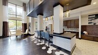 Located at 910 Deerfield Crossing Dr in Alpharetta, GA, this single-floor space offers a thoughtfully designed open-concept layout that seamlessly blends a fully equipped kitchen with a generous living area. Soaring double-height ceilings amplify the sense of openness throughout, while large floor-to-ceiling windows invite abundant natural light and provide views of the surrounding greenery.

The kitchen is a standout feature, appointed with rich dark-wood cabinetry, sleek dark quartz countertops, a mosaic tile backsplash, and stainless steel appliances. Distinctive large-format hexagonal tile flooring anchors the kitchen zone, transitioning into warm, wide-plank hardwood flooring in the adjoining areas. A generously sized island with white subway tile detailing and bar seating, illuminated by a series of elegant gold pendant lights, serves as a central gathering point.

The living area is defined by a bold design palette — dark wainscoting, crisp white upper walls, and structural columns that add architectural interest. Comfortable seating arrangements and curated wall art contribute to a polished yet welcoming atmosphere. A wall-mounted display screen adds a functional element to the space. Together, these areas create a cohesive, upscale environment well-suited for social gatherings and community interaction.