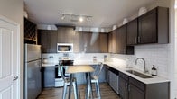 This single-floor 1-bedroom, 1-bathroom residence offers a thoughtfully designed layout with modern finishes that create a cohesive and inviting atmosphere. The kitchen is a standout feature, showcasing rich dark-toned cabinetry that extends to the ceiling for generous storage capacity. Light quartz-style countertops provide a striking contrast against the dark cabinets, while a classic white subway tile backsplash adds a clean, timeless aesthetic. Stainless steel appliances, including a range, microwave, and dishwasher, provide a sleek, contemporary feel. Track lighting overhead ensures the kitchen is well-lit and functional. A compact kitchen island with two industrial-style metal bar stools creates a casual dining or gathering space within the kitchen area. The wood-look flooring runs throughout, adding warmth and continuity to the space. The overall design balances dark and light elements to create a modern yet welcoming environment, making this residence a comfortable and stylish place to call home.