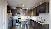 This single-floor 1-bedroom, 1-bathroom residence offers a thoughtfully designed layout with modern finishes that create a cohesive and inviting atmosphere. The kitchen is a standout feature, showcasing rich dark-toned cabinetry that extends to the ceiling for generous storage capacity. Light quartz-style countertops provide a striking contrast against the dark cabinets, while a classic white subway tile backsplash adds a clean, timeless aesthetic. Stainless steel appliances, including a range, microwave, and dishwasher, provide a sleek, contemporary feel. Track lighting overhead ensures the kitchen is well-lit and functional. A compact kitchen island with two industrial-style metal bar stools creates a casual dining or gathering space within the kitchen area. The wood-look flooring runs throughout, adding warmth and continuity to the space. The overall design balances dark and light elements to create a modern yet welcoming environment, making this residence a comfortable and stylish place to call home.
