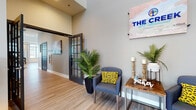 The Creek at Cottonwood Apartment Homes leasing office is a thoughtfully designed, single-floor community space that makes a strong first impression. The grand entrance features soaring ceilings, floor-to-ceiling glass-paneled doors, and light wood-style flooring that flows throughout the entire space, creating a warm and cohesive aesthetic.

The welcoming lobby greets visitors with comfortable seating, tropical greenery, and branded signage that sets an inviting tone. French-style interior doors with dark frames add an elegant architectural detail that carries through multiple areas of the space.

A central lounge area provides a relaxed setting with plush seating, a wall-mounted television framed by decorative geometric paneling, and carefully curated décor. Adjacent to the lounge, a bright and open gathering room benefits from an abundance of natural light streaming through large windows fitted with patterned drapes, offering views of the surrounding neighborhood. This area accommodates multiple seating arrangements, including a communal dining table, making it ideal for resident events.

The kitchen is well-appointed with dark cabinetry, light countertops, full appliances, and pendant lighting overhead. A large island with bar seating serves as a natural gathering point. A cozy nook with a bistro-style table and colorful artwork provides an additional informal seating option. Together, these spaces reflect a modern, community-oriented design built for comfort and functionality.