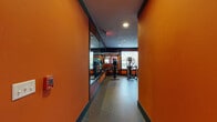 This fitness center is designed with a bold, energetic aesthetic defined by vivid orange walls accented with dark charcoal trim, creating a motivating atmosphere throughout. The space is organized into distinct functional zones connected by a corridor entrance flanked by full-length mirrors that visually expand the area upon entry.

The cardio zone is the largest section, housing an extensive row of treadmills equipped with individual screens, alongside a lineup of elliptical trainers and stationary bikes. Large windows along the walls allow natural light to filter in, offering views of surrounding greenery and contributing to an open, airy feel. Mirrors are strategically placed to further enhance the sense of space.

A separate functional training area provides open floor space finished with durable rubber flooring, ideal for stretching and bodyweight exercises. This zone includes a dumbbell rack, medicine balls, exercise mats, and wall-mounted storage for accessories, supporting a variety of workout routines.

Recessed ceiling lighting ensures consistent illumination throughout, complemented by a ceiling fan in the cardio area for added ventilation. The rubber flooring, used consistently across all zones, provides both durability and safety. Overall, the space is thoughtfully laid out to accommodate a full range of fitness activities in a cohesive, well-appointed environment.
