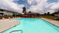 Located at 160 Peaceful Ln in Sanford, North Carolina, this single-story community amenity space centers around a generously sized outdoor swimming pool with clear, turquoise water. The pool is surrounded by a wide concrete sun deck equipped with multiple lounge chairs, providing ample space for relaxing and sunbathing. A dedicated pool house structure sits poolside, offering restroom and utility access for guests. Framing the deck is a covered porch area with a warm wood-paneled ceiling, ceiling fan, and classic dark-stained rocking chairs — creating a shaded retreat ideal for unwinding while overlooking the pool. The porch connects to an interior space through multiple white-framed French doors with glass pane detailing, lending an open and airy feel to the transition between indoor and outdoor areas. Mature pine trees and well-maintained greenery form a natural backdrop around the pool area, enhancing the serene, park-like atmosphere. Multi-story residential buildings are visible in the background, contextualizing this space as a central community gathering hub. The overall setting is open, bright, and inviting, designed to encourage outdoor recreation and social activity in a well-appointed, landscaped environment.