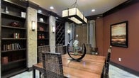 This elegantly designed single-floor space makes a striking impression through its carefully curated interior aesthetic. The dining area serves as a central showpiece, anchored by a solid wood dining table surrounded by woven rattan chairs that add organic texture to the rich, dark palette. Overhead, a rectangular pendant light fixture with a warm diffused glow provides both functional illumination and a bold architectural statement.

Flanking the space are stacked stone columns that lend a natural, earthy contrast to the dark wood built-in shelving and cabinetry. These built-ins incorporate a diamond-pattern wine rack, offering both practical storage and a visually dynamic design element. Wall-mounted sconces complement the recessed ceiling lighting, creating a layered and intimate ambiance throughout.

Deep, warm-toned walls — rendered in rich terracotta hues — enhance the cozy yet sophisticated atmosphere. Roman-style window shades in neutral tones filter natural light softly into the room, while decorative drapery panels add depth and texture to the windows. A sculptural centerpiece on the dining table adds an artistic finishing touch. Overall, this space exudes a refined, lodge-inspired warmth with a distinctly upscale character.