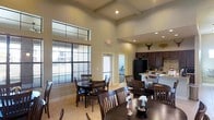 This single-story space serves as a communal gathering area, thoughtfully designed to accommodate dining and socializing on a generous scale. The room immediately draws attention upward with its soaring ceilings, which are accentuated by coffered detailing and recessed lighting that creates a bright, welcoming atmosphere throughout. Clerestory windows positioned high on the walls allow natural light to filter in while maintaining privacy, complementing the large lower windows fitted with horizontal blinds that offer views of the surrounding exterior.

The dining area is well-appointed with multiple dark wood tables and matching chairs arranged to encourage both intimate gatherings and larger group seating. The tile flooring throughout provides a durable and clean foundation, consistent with a high-traffic communal environment.

Adjacent to the dining area, a full kitchen is visible, featuring dark cabinetry, a large refrigerator, stone-look countertops, and a tile backsplash. The kitchen island provides additional workspace and informal seating. A decorative longhorn mount adds a regional character to the space.

Beyond the main area, a doorway hints at additional rooms, potentially a fitness or activity area, further expanding the functional versatility of this well-equipped shared space.