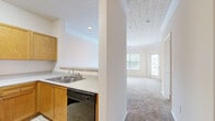 This thoughtfully laid-out single-floor residence offers a comfortable and functional living environment across its well-defined rooms. The combined kitchen and living area forms the heart of the home, with the kitchen featuring warm honey-toned oak cabinetry, light-colored countertops, black appliances including a range and dishwasher, and tile flooring. A half-wall pass-through connects the kitchen to the living area, creating an open, airy feel. The living room is generously sized, finished with plush wall-to-wall carpeting, crown molding, a textured ceiling, and multiple windows that allow natural light to fill the space. A sliding glass door provides access to an outdoor balcony area with views of surrounding greenery. The spacious bedroom continues the warm, neutral color palette with soft carpeting, a ceiling fan with light fixture, and a window overlooking the balcony and wooded surroundings. The bedroom includes direct access to both a dedicated closet and the full bathroom. The bathroom is appointed with an oak vanity, ample counter space, a large wall mirror, and tile flooring. A hallway connects the main living areas, completing this cohesive and efficiently designed floor plan.