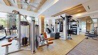This fitness center combines contemporary design with warm, natural materials to create a spacious and inviting workout environment. Exposed wood beams span the ceiling throughout, complemented by wood-clad columns that add architectural character and a sense of organic warmth to the space. Large-format windows and floor-to-ceiling glass panels flood the interior with abundant natural light and provide views of surrounding greenery, enhancing the overall atmosphere.

The facility is thoughtfully organized into several distinct workout zones. A dedicated cardio area features a row of treadmills, ellipticals, and stair climbers positioned along the expansive windows. A functional training zone is equipped with TRX suspension systems, battle ropes, a overhead rig, kettlebells, medicine balls, and a heavy punching bag. The strength training area includes Precor cable machines, a power rack with benches, and a comprehensive free weight selection. A mirrored studio space with a ballet barre and open floor area accommodates stretching and group exercise. Cork and hardwood flooring, large ceiling fans, and steel-framed glass doors complete the polished, high-end aesthetic.