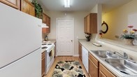 Located on Kings Ridge Drive in Charlotte, NC, this single-story residence offers a thoughtfully arranged floor plan that balances comfort and functionality. The layout, visible in the 3D overview, includes two bedrooms, two bathrooms, a living room, a dining area, and a galley-style kitchen, all connected by a straightforward, accessible flow.

The living room features neutral beige walls, carpet flooring, and multiple windows dressed with layered curtains and blinds that allow generous natural light. The adjoining dining area accommodates a round table and transitions seamlessly toward the kitchen.

The galley kitchen is lined on both sides with warm wood cabinetry and light countertops, and includes white appliances—a refrigerator, electric range, and dishwasher—alongside a double sink. Wood-look flooring runs throughout the kitchen.

Both bedrooms offer carpeted floors, natural light from windows with blinds and curtains, and ample wall space. Each is served by its own full bathroom, each featuring a single-sink vanity with wood cabinetry, a tub-shower combination with a curtain rod, and wood-look flooring. The overall aesthetic throughout the residence is warm and neutral, with consistent finishes tying the space together.