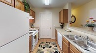 Located on Kings Ridge Drive in Charlotte, NC, this single-story residence offers a thoughtfully arranged floor plan that balances comfort and functionality. The layout, visible in the 3D overview, includes two bedrooms, two bathrooms, a living room, a dining area, and a galley-style kitchen, all connected by a straightforward, accessible flow.

The living room features neutral beige walls, carpet flooring, and multiple windows dressed with layered curtains and blinds that allow generous natural light. The adjoining dining area accommodates a round table and transitions seamlessly toward the kitchen.

The galley kitchen is lined on both sides with warm wood cabinetry and light countertops, and includes white appliances—a refrigerator, electric range, and dishwasher—alongside a double sink. Wood-look flooring runs throughout the kitchen.

Both bedrooms offer carpeted floors, natural light from windows with blinds and curtains, and ample wall space. Each is served by its own full bathroom, each featuring a single-sink vanity with wood cabinetry, a tub-shower combination with a curtain rod, and wood-look flooring. The overall aesthetic throughout the residence is warm and neutral, with consistent finishes tying the space together.