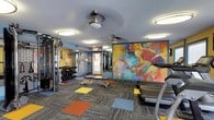 The Parkway Square fitness center is a thoughtfully designed workout facility that combines functionality with an energetic, artistic atmosphere. The space is finished with a multi-toned carpet featuring bold color-block accents in yellow, red, blue, and orange, adding a dynamic visual element to the floor throughout. Walls are painted in a warm yellow along one side and a cool blue-gray on the other, creating a lively contrast that contributes to the room's motivating ambiance.

A striking large-scale abstract mural of a tennis player dominates one wall, serving as a bold focal point alongside additional framed artwork displayed throughout. Full-length mirrors line a portion of the wall, visually expanding the space and aiding users during workouts. Natural light enters through several windows fitted with blinds, complemented by flush-mount and pendant ceiling fixtures that provide consistent overhead lighting.

The cardio zone is equipped with treadmills and elliptical machines, each accompanied by wall-mounted televisions for entertainment. The strength area includes a dumbbell rack, an adjustable weight bench, cable machines, and additional resistance equipment. Water stations and sanitation supplies are conveniently positioned within the space, ensuring a clean and accessible environment for all users.