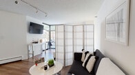 This single-floor studio unit is designed with an open-concept layout that maximizes every inch of available space. A striking six-panel Japanese-style shoji folding screen serves as the primary room divider, elegantly separating the sleeping area from the living space while maintaining an airy, cohesive feel throughout. The living area features warm hardwood-style flooring, a wall-mounted flat-screen TV, and a contemporary sofa paired with a round white coffee table, creating a comfortable and stylish lounge space. Track lighting runs along the textured ceiling, providing adjustable illumination across the unit. A corner window arrangement floods the workspace and lounge area with natural light and outdoor views of surrounding greenery. The sleeping area is defined by the folding screen partition and includes a large mirror, adding depth to the room. The kitchen is efficiently laid out in an L-shape, equipped with black appliances including a range, microwave, and dishwasher, complemented by dark lower cabinetry and white upper cabinets. The bathroom features classic black-and-white mosaic tile flooring, a dark wood vanity, and a well-lit mirror cabinet, rounding out this thoughtfully appointed space.