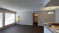 This single-story space in Dallas, Texas offers a straightforward, functional layout with a clean and neutral aesthetic throughout. The main living area features dark hardwood-style flooring that provides a rich contrast against the light gray walls, creating a grounded and cohesive look. A textured popcorn ceiling adds a classic residential character to the space, while the HVAC vent integrated into the ceiling ensures climate comfort year-round.

Natural light enters through a window along the left wall, fitted with horizontal blinds that allow for adjustable light control. The open sightlines connect the living area to an adjacent room visible through a doorway, suggesting a functional flow between spaces. To the right, a partial view of a countertop with a granite or stone surface hints at a kitchen or bar area, indicating a convenient proximity between living and dining or cooking zones.

The neutral color palette and minimal detailing throughout give the space a versatile canvas suitable for a variety of interior styles. White baseboards provide a clean transition between the walls and flooring, reinforcing the tidy, well-maintained character of this Dallas residence.
