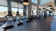 This single-floor fitness center is thoughtfully designed to accommodate a wide range of workout routines within a modern, open layout. The space is divided into distinct functional zones, each tailored to specific training needs. Large windows line the walls throughout, flooding the interior with abundant natural light and offering views of the surrounding neighborhood, creating an energizing atmosphere for exercise.

The cardio area features a row of treadmills and elliptical cross-trainers positioned along the windowed walls, allowing users to enjoy the outdoor scenery while working out. A dedicated cycling studio houses multiple Matrix spin bikes arranged beneath bold, oversized pendant lights, with a wall-mounted flat-screen TV providing entertainment or guided class content. Full-length mirrors along one wall visually expand the space.

A separate strength training zone includes a dumbbell rack with a range of free weights, adjustable benches, and cable-based strength machines, providing options for both beginners and experienced athletes. Durable rubber flooring runs throughout, offering safety and comfort underfoot. The facility also includes restrooms conveniently accessible from the main corridor, which is accented with contemporary artwork, completing the polished, well-appointed aesthetic.