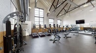 This fitness center occupies a generously sized single-floor space that retains the character of its industrial origins. Soaring vaulted ceilings with exposed white-painted rafters and dark structural steel trusses create an open, airy atmosphere overhead, while large multi-pane windows line the walls, flooding the interior with natural light and offering views of the surrounding exterior. Warm-toned brick tile wainscoting runs along the lower portion of the walls, adding a classic industrial aesthetic that contrasts with the clean white upper walls.

The flooring throughout consists of durable rubber gym matting, practical for both heavy equipment and high-impact workouts. The cardio zone features a row of treadmills, elliptical trainers, upright and recumbent bikes, all equipped with individual display screens. A large flat-screen television is mounted on the wall, providing entertainment for users.

The strength training area is well-appointed with multiple cable and multi-function weight machines, adjustable benches, and a dedicated dumbbell rack. A partitioned section with frosted glass panel dividers offers a more private training environment for additional strength machines. Exposed ductwork and pendant lighting fixtures hang from the ceiling, reinforcing the industrial character of the space while maintaining a bright, functional workout environment.
