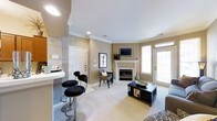 This single-floor residence offers a thoughtfully designed open-concept layout that seamlessly connects the living and dining areas. The living room serves as the focal point of the space, anchored by a classic white-mantled fireplace with a wall-mounted television above, creating a warm and inviting atmosphere. Crown molding throughout adds an elegant architectural detail, while recessed lighting provides even, ambient illumination across the neutral-toned walls and plush carpeted floors.

Natural light fills the space generously through large windows and a glass-paneled door, enhancing the bright and airy feel of the interior. The seating area is complemented by a dark wood coffee table and an accent chair, offering a comfortable and functional arrangement. Adjacent to the living area, a bar-style counter with sleek black stools defines a casual dining nook, ideal for quick meals or entertaining. The entryway features a solid paneled front door with clean white trim, contributing to the space's polished, cohesive aesthetic. Overall, the residence combines classic design elements with modern touches to create a warm, livable environment.