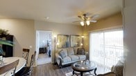 This single-floor residence offers a thoughtfully designed layout that balances comfort and functionality. The living area serves as the heart of the space, featuring warm hardwood-style flooring, neutral wall tones, and a ceiling fan with integrated lighting that enhances airflow and ambiance. A large sliding glass door floods the room with natural light and provides direct access to a private outdoor area, complemented by vertical blinds for adjustable light control. The living space is generously sized to accommodate a full seating arrangement alongside accent furniture, creating an inviting atmosphere for relaxing or entertaining. Abstract wall art adds a contemporary touch to the otherwise classic décor. An open doorway connects the living area directly to the bedroom, which continues the warm, neutral color palette and features carpeted flooring for added comfort. The bedroom is spacious enough to accommodate a full bedroom set, including a bed frame, nightstands, and additional accent pieces. Throughout the residence, the design maintains a cohesive aesthetic that is both welcoming and practical, making effective use of the available space across the single floor.