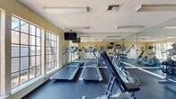 This fitness center is a well-appointed, single-room exercise facility designed to offer a comprehensive workout experience within a compact footprint. The space features warm yellow walls, a textured ceiling, and durable dark rubber flooring that provides both safety and functionality for various exercise activities.

Natural light flows generously through multiple large windows fitted with decorative grilles, offering views of the surrounding outdoor area and creating an open, airy atmosphere. Full-length mirrors line one wall, effectively doubling the perceived size of the room while allowing users to monitor their form during workouts.

The facility is thoughtfully equipped with a lineup of cardio machines, including multiple treadmills and an elliptical trainer, positioned to take advantage of the natural light from the windows. A wall-mounted flat-screen television provides entertainment during cardio sessions. The strength training area includes a dual cable machine, an adjustable workout bench, a rack of dumbbells in varied weights, and a medicine ball storage tower with an assortment of colorful exercise balls. Overhead fluorescent lighting ensures consistent illumination throughout. A posted fitness center rules board near the entrance reflects the organized, community-oriented nature of the space.