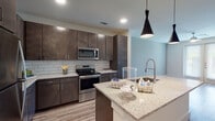 This well-appointed single-floor residence offers a thoughtfully designed open-concept layout that seamlessly connects the kitchen and living areas. The kitchen is a standout feature, showcasing rich dark-espresso cabinetry, light granite countertops, and a full suite of stainless steel appliances including a refrigerator, electric range, and over-the-range microwave. A classic subway tile backsplash adds texture and visual interest, while a central island with a modern pendant light above provides additional prep space and a natural gathering point. Wood-look plank flooring runs throughout the main living areas, lending warmth and continuity to the space. The living area is bright and open, enhanced by natural light from French doors that lead to an exterior balcony, and is completed with a ceiling fan. The bedroom offers plush carpeting, a ceiling fan, and a window that invites natural light. The bathroom is clean and functional, featuring a granite-topped vanity with dark cabinetry, wood-look tile flooring, and access to a built-in walk-in closet with dedicated shelving. The overall aesthetic is contemporary and cohesive, balancing neutral tones with modern fixtures for a comfortable, move-in-ready feel.
