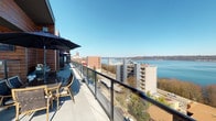 This expansive rooftop terrace offers an exceptional outdoor living experience defined by its commanding, unobstructed views. The space is finished with large-format light-toned pavers underfoot and bordered by sleek glass-panel railings that preserve sightlines in every direction. Warm, richly toned wood cladding on the building's exterior facade adds a natural contrast to the modern architectural design and creates an inviting backdrop throughout the terrace.

Multiple distinct seating areas are thoughtfully arranged along the terrace. A generous dining setup anchored by a large market umbrella provides a shaded space for outdoor gatherings, complemented by a high-top bistro table and bar seating positioned to take full advantage of the views. A lounging area features contemporary hanging egg chairs and sleek sun loungers, ideal for relaxing while soaking in the surroundings. Mounted televisions are integrated into covered sections of the exterior wall, extending the functionality of the space.

The views from this terrace are truly a standout feature. Sweeping vistas encompass a broad waterway with active harbor activity, a lush park below, a vibrant urban cityscape, and a prominent snow-capped mountain peak visible on the horizon — all under expansive open skies. The combination of thoughtful design, high-quality finishes, and breathtaking panoramic scenery makes this terrace a remarkable outdoor destination.