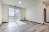 This thoughtfully designed single-floor unit offers a functional and modern layout across six distinct spaces, including a kitchen, living room, bedroom, bathroom, laundry room, and closet. The open-concept kitchen is a standout feature, showcasing rich medium-toned wood cabinetry, granite countertops, and a full suite of stainless steel appliances including a refrigerator, electric range, microwave, and dishwasher. A central kitchen island with a matching granite surface provides additional prep space and casual seating potential. A sleek multi-pendant black light fixture adds a contemporary touch overhead. Light wood-style flooring flows throughout the main living areas, lending warmth and continuity to the space. The living room is bright and airy, benefiting from a large sliding glass door that brings in natural light and outdoor views. The bedroom is generously sized, finished with plush carpet, a ceiling fan with light, and a wide window overlooking the surrounding area, complemented by an adjacent closet. The bathroom features a full tub-shower combination with chrome fixtures and a granite-topped vanity. A dedicated laundry room with a full-size washer and dryer and overhead shelving adds exceptional everyday convenience to this well-rounded unit.